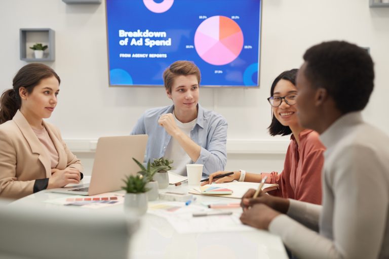 Multi-ethnic group of young business people brainstorming ideas in conference room, focus on smiling man heading meeting table